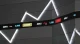 Stock ticker board showing company symbols and numeric values on a dark wall, with large illuminated zigzag light fixtures above.