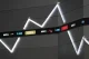 Stock ticker board showing company symbols and numeric values on a dark wall, with large illuminated zigzag light fixtures above.