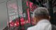 Person stands in front of a glass wall showing stock market tickers and red digital charts, as they read the numbers.