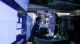 A man in a dark shirt stands at a desk in a dim, blue-lit control room, monitoring multiple computer screens and dashboards.