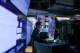 A man in a dark shirt stands at a desk in a dim, blue-lit control room, monitoring multiple computer screens and dashboards.