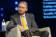 Panel speaker: man in a gray checkered suit and orange tie, seated on stage with a headset mic, blue lights in the background.