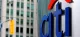 Close-up of the blue Citi logo with a red arc on a metal sign, with a blurred city street in the background.