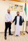 Two men shake hands in an office next to a United Arab Emirates flag and framed portraits on the wall, presenting a formal meeting scene.