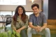 Two smiling coworkers sit closely on a tan couch in a bright, modern office.