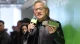 Man with gray hair and glasses speaks into a lime-green microphone at an event, holding a green prop.