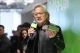 Man with gray hair and glasses speaks into a lime-green microphone at an event, holding a green prop.