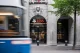 Blurred blue city bus passes in front of a stone UBS bank entrance with arched doorway and a carved sculpted face above the arch, flags visible above.