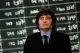 Man in a dark suit and blue shirt stands in front of a blurred stock market board with numbers.