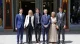 Six people in business attire stand arm-in-arm outside a storefront, posing for a formal group photo in daytime sun.