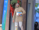 Man in a tan coat and checked pants stands at a doorway, holding magazines and wearing sunglasses and a smartwatch next to a festive wreath and blue mural.