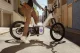 Person in a denim jacket and brown pants stands beside a white electric bike with purple accents on a city street.
