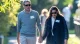 Couple wearing sunglasses strolls along a sunlit park path, holding hands and wearing name badges.