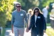 Couple wearing sunglasses strolls along a sunlit park path, holding hands and wearing name badges.