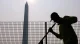 Construction worker in a high-visibility jacket leaning on a fence with the Washington Monument in the background under a hazy sun (silhouette).