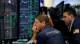 Two stock traders in blue jackets study multiple trading screens filled with candlestick charts and numbers, one resting his chin in thought as he watches the data.