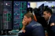 Two stock traders in blue jackets study multiple trading screens filled with candlestick charts and numbers, one resting his chin in thought as he watches the data.